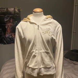 Vit hoodie från Juicy Couture - Vit hoodie med dragkedja och huva från Juicy Couture. Broderad logga med krona och texten 'Juicy Girl' i guld på både fram- och baksidan. Mjuk och skön, perfekt för en avslappnad stil.