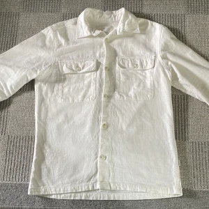 Vit overshirt från Jack & Jones - Säljer en stilren vit overshirt från Jack & Jones. Skjortan har två bröstfickor med knappar och klassisk krage. Perfekt att bära öppen eller stängd för en clean look.