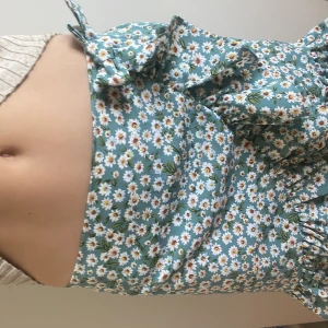 Blommig omlottkjol i grönt - Säljer en söt kort omlottkjol med volangkant och blommigt mönster i vitt och gult på grön botten. Kjolen har knyt i midjan och är perfekt för dig som gillar en feminin och somrig stil.