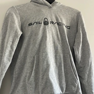 Grå hoodie från Sail Racing - Säljer en ljusgrå hoodie från Sail Racing med svart logga på bröstet och en mindre logga på huvan. Tröjan har känguruficka och är tillverkad i mjukt bomullsmaterial. Perfekt för en avslappnad stil.