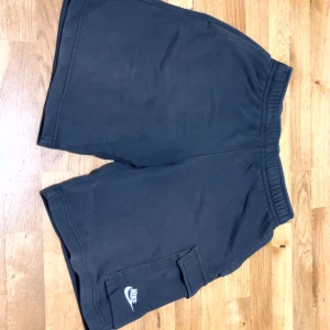 Svarta shorts från Nike med cargoficka - Svarta shorts från Nike med elastisk midja och en stor cargoficka på sidan. Klassisk Nike-logga i vitt på fickan. Perfekta för träning eller avslappnade dagar. Tillverkade i mjukt material för extra komfort.