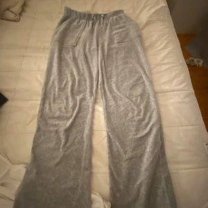 Grå mjukisbyxor från Juicy Couture - Supermjuka grå mjukisbyxor från Juicy Couture med bred resår i midjan och dragsko. Byxorna har raka ben och två fickor bak med lock och silvriga knappar. Perfekta för en avslappnad stil.