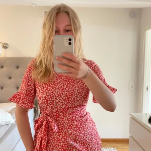 Röd blommig klänning från Vero Moda - Söt röd kortklänning från Vero Moda med vitt blommönster. Klänningen har korta ärmar, volangdetaljer och knyt i midjan för en fin passform. Perfekt för sommarens alla tillfällen!