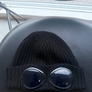 Svart ribbstickad mössa med glasögondetalj från C.P. Company - Cp company mössa. Den är i bra skick och köpt från butiken: Jupiter för ca 1599kr. 