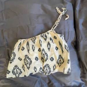 Mönstrad offshoulder topp med knytband - Säljer en beige och svart offshoulder topp med trendigt mönster och flätat knytband på ena axeln. Tunt, luftigt material som passar perfekt till sommaren. Snygg och unik detalj med det grafiska mönstret. Från Zara, lite missfärgad som ni ser på andra bilden men syns knappt