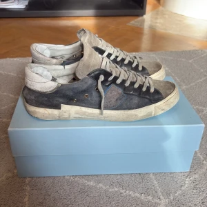 Philippe Model sneakers i grå mocka - Säljer ett par Philippe Model sneakers i grå och beige mocka med vita detaljer. Skorna har snörning och en platt sula. Klassisk design med rund tå och diskret logga på sidan. Perfekta för dig som gillar stilrena och bekväma sneakers. Box och tillbehör medföljer. Skorna är laggade inuti. Men skicket är väl ändå 6/10 ungefär. Det är storlek 41.