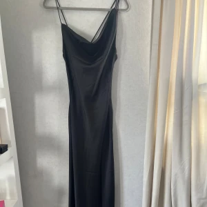 Svart långklänning med tunna axelband från Boohoo - Säljer en svart långklänning/balklänning från Boohoo med tunna axelband och draperad urringning. Klänningen har en elegant, slimmad passform och är tillverkad i ett glansigt, mjukt material som ger en lyxig känsla.Aldrig använd,endast testad säljer den för att jag köpte en ny klänning. Storlek 38 men jag som oftas har storlek 36 så passar den perfekt.