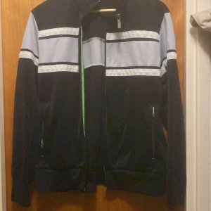 Svart track jacket från Hugo Boss med vita ränder - Säljer en svart track jacket från Hugo Boss med vita och ljusgrå horisontella ränder över bröstet och armarna. Jackan har dragkedja framtill och två fickor med dragkedja. Klassisk sportig look med ribbade muddar.