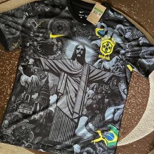 Brasilien fotbollströja med Kristusstatyn - Unik Brasilien fotbollströja från Nike med svart bas och detaljerat grafiskt tryck av Kristusstatyn, gula Nike-loggan och Brasilien-emblem på bröstet. Kortärmad modell med rund hals och inslag av blått och gult i designen.
