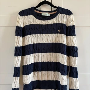 Randig kabelstickad tröja från Morris - Säljer en kabelstickad tröja från Morris i marinblått och vitt med breda ränder. Tröjan har rund halsringning och lång ärm, samt en liten broderad logga på bröstet. Perfekt för en klassisk och stilren look. Sitter mer som en S.