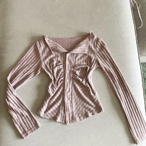 Rosa ribbad croppad kofta med knappar - Säljer en ljusrosa ribbad kofta med långa ärmar och knappar framtill. Tröjan är croppad och har en bred krage. Perfekt för dig som gillar en trendig och feminin stil.