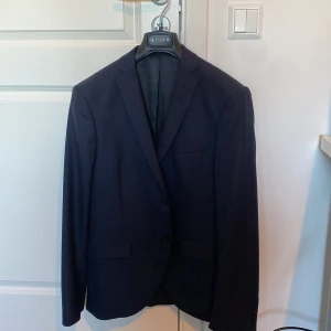 Mörkblå kavaj från Tiger of Sweden - Säljer en stilren mörkblå kavaj från Tiger of Sweden i klassisk modell. Kavajen har två knappar framtill, smal passform och är tillverkad i en blandning av polyester och ull. Perfekt för student eller festliga tillfällen.