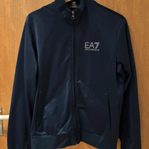 Mörkblå kofta från EA7 Emporio Armani - Snygg mörkblå kofta från EA7 Emporio Armani med dragkedja framtill och logga på bröstet. Jackan har hög krage, ribbade muddar och lång ärm. Perfekt för en sportig och stilren look.