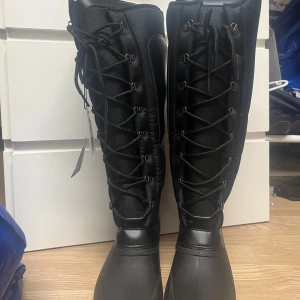 Svarta ridstövlar med snörning och dragkedja - Säljer ett par höga svarta ridstövlar med snörning framtill och dragkedja på baksidan. Stövlarna är helt nya med prislapp.. Perfekta för dig som vill ha både stil och funktion i stallet.  Ena dragkedjan dvs ” dragkroken ” man tar tag i har gått av från ena sidan på högra stöveln. Kan gå att lösa med antingen ny drag grejs eller så kanske det behövs bytas dragkedja helt.  Normal i storleken men långa. Vid fler bilder eller frågor, kontakta mig 🤗🙋🏼‍♀️