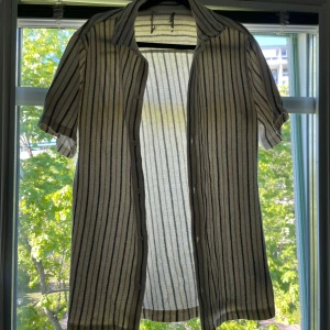 Randig kortärmad skjorta från 157 - Säljer en luftig och stilren kortärmad skjorta från 157 med svarta och grå ränder på vit botten. Skjortan har klassisk krage och knappar framtill. Perfekt för varma dagar och enkel att matcha med jeans eller shorts. Klippt av lappen i mitten då den skavde i nacken som man kan se på första bilden.