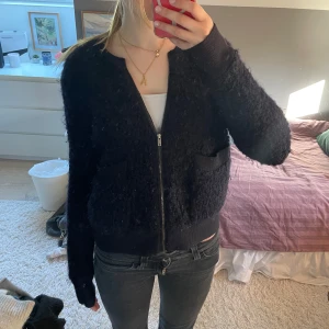 Svart stickad bomberjacka med dragkedja - Mysig svart stickad bomberjacka med dragkedja framtill och två fickor. Jackan har ribbade muddar vid ärmslut och midja, och en klassisk rund halsringning. Perfekt för lager-på-lager och enkel att matcha.