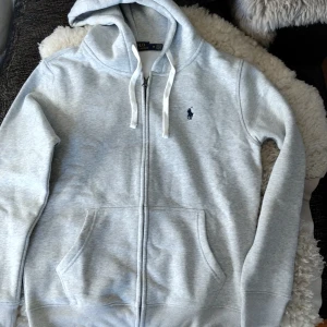 Grå hoodie från Polo Ralph Lauren - Säljer en ljusgrå hoodie från Polo Ralph Lauren med dragkedja och vit dragsko i huvan. Tröjan har två fickor framtill och den klassiska broderade loggan på bröstet. Perfekt för en avslappnad stil.