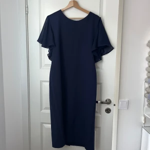 Marinblå klänning med volangärmar - Säljer en stilren marinblå klänning med vackra volangärmar och rund halsringning. Klänningen har en rak passform och stängs med dragkedja i ryggen. Perfekt för dig som gillar enkel men elegant stil.