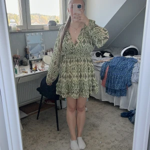 Grön mönstrad kortklänning med volang - Säljer en kort grön klänning med svart och vit mönster, v-ringning och långa, luftiga ärmar. Klänningen har smock i midjan och volangdetaljer nertill. Perfekt för sommarens alla tillfällen!