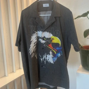 Rhude kortarmad skjorta - Säljer en grå skjorta från Rhude med ett stort färgstarkt tryck av en örn på framsidan. Skjortan har korta ärmar, klassisk krage och knäppning framtill. Perfekt statement-plagg för dig som vill sticka ut.