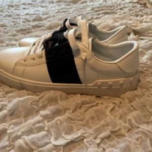 Valentino sneakers med svart detalj - Säljer ett par vita Valentino sneakers med en bred svart rem över mitten och nitar längs sulan. Skorna har rund tå och platt sula, samt klassisk snörning. Perfekta för dig som gillar stilrena men ändå unika skor. I princip nya, sällan använda.  