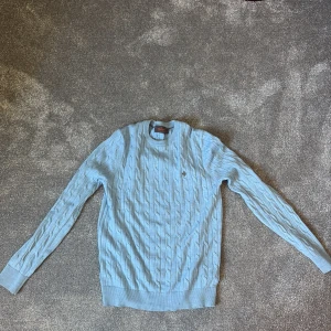 Morris - Säljer en ljusblå kabelstickad tröja från Morris med rund halsringning och broderad logga på bröstet. Tröjan har långa ärmar och ribbade muddar vid ärmslut och nederkant. Perfekt för lager-på-lager under kyliga dagar. Merin ull som anpassar sig efter väder. Men den har ett litet hål.