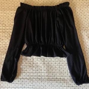Svart offshoulder blus med volang - Säljer en svart offshoulder blus med långa ärmar och volang nertill. Blusen har resår vid axlar och midja samt ett diskret prickigt mönster i tyget. Blusen har knappt använts och är i mycket bra skick.