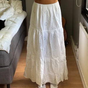Vit långkjol med broderade blommor - Luftig och somrig vit långkjol med broderade blommor och hålmönster från Gina Tricot. Kjolen har flera volanger och en lätt utsvängd passform som ger en bohemisk känsla. Aldrig använd, endast provad med prislapp kvar. 300 kr