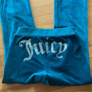 Turkosa byxor från Juicy Couture - Säljer ett par turkosa velourbyxor från Juicy Couture med strassdetaljer och stor Juicy-text bak. Byxorna har bred resår i midjan och klassisk mjukisbyxmodell. Perfekta för en avslappnad och trendig look.