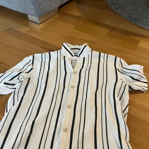 Vit och svart randig skjorta från Zara - Säljer en snygg vit skjorta från Zara med svarta ränder och korta ärmar. Skjortan har knappar framtill och är tillverkad i ett lätt linnematerial, perfekt för varma dagar. Stilig ståkrage ger en modern touch.