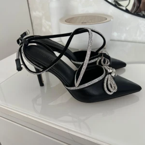 Svarta klackskor med rosett  - Säljer ett par svarta klackskor med spetsig tå och tunna remmar dekorerade med glittrande strass. Skorna har en elegant rosettdetalj framtill med pärlor och stenar. Hör av er vid intresse🥰. 