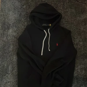 Svart hoodie från Polo Ralph Lauren - Svart hoodie från Polo Ralph Lauren med vit dragsko och klassisk röd logga på bröstet. Mjuk och bekväm, perfekt för en avslappnad stil. Passar dig som gillar stilrena och tidlösa plagg.
