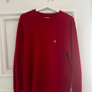Röd långärmad tröja från Polo Ralph Lauren - Röd långärmad tröja från Polo Ralph Lauren med rund halsringning och vit broderad logga på bröstet. Tillverkad i ett mjukt material som passar perfekt för en avslappnad stil.