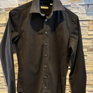 Svart skjorta från burberry - Stilren svart skjorta från burberry med klassisk krage och knäppning framtill. Skjortan har långa ärmar och diskret logga broderad på bröstet. Perfekt för dig som vill ha en tidlös och elegant look.