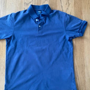 Blå pikétröja Uniqlo - Säljer en klassisk blå pikétröja från Uniqlo med krage och två knappar framtill. Tröjan är kortärmad och har en stilren design som passar perfekt till sommaren. Tillverkad i mjuk bomull för en bekväm känsla.