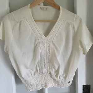 Vit blus med spetsdetaljer - Säljer en söt vit blus med v-ringning och fina spetsdetaljer framtill. Blusen har korta ärmar och smockad nederkant som ger en snygg passform. 