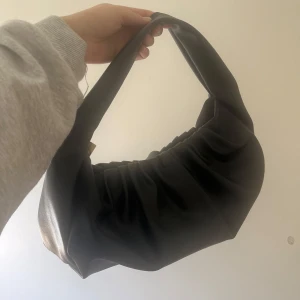 Svart axelväska i syntetmaterial - Trendig svart stor axelväska i mjukt syntetmaterial med rynkade detaljer och dragkedja i guld. Perfekt storlek för det viktigaste och enkel att bära över axeln. Snygg och stilren design som passar till många outfits. Får plats mycket i den. 