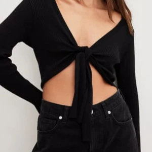 Svart ribbad croppad topp med knyt - Trendig svart croppad topp med lång ärm och djup v-ringning. Toppen är ribbad och har en snygg knytning framtill som ger en fin look.