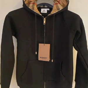 Svart hoodie från Burberry med klassiskt rutigt foder - Säljer en svart hoodie från Burberry med dragkedja och huva. Insidan av huvan har det ikoniska Burberry-rutiga fodret. Tröjan har fickor framtill och guldfärgade detaljer på snöret. Perfekt för dig som vill ha en lyxig och stilren look.