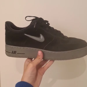 Svarta Nike Air Force 1 sneakers - Säljer ett par svarta Nike Air Force 1 sneakers med grå sula och grå swoosh. Skorna har snörning och är tillverkade i mocka med klassisk rund tå och platt sula. Perfekta för dig som gillar stilrena och tidlösa sneakers. Köpte dom för 1700 och är knappt använd 