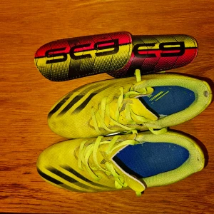 Gula fotbollsskor med svarta detaljer - Säljer ett par gula Adidas fotbollsskor med svarta ränder och blå innersula. Skorna har snörning och är designade för att ge bra grepp på planen. Perfekta för dig som vill sticka ut på fotbollsplanen!