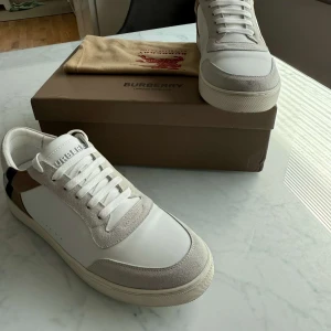 Burberry skor  - Snygga vita sneakers från Burberry med grå mockadetaljer och klassiskt rutigt mönster på hälen. Skorna har snörning och rund tå, samt en platt sula. Perfekta för dig som vill ha en stilren och lyxig look.