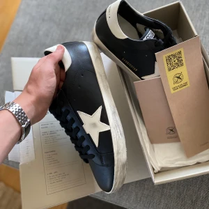 Golden Goose Super-Star sneakers i svart och vitt - Golden Goose Super-Star sneakers i svart skinn med vit stjärndetalj på sidan och vit häl. Klassisk rund tå och platt sula. Svarta skosnören och GGDB-logga på plösen. Perfekta för en cool och avslappnad stil.