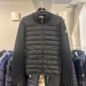 Moncler Down Cardigan S NYSKICK - Snygg svart cardigan jacka från Moncler med quiltad framsida och ärmar i stickad bomull. Jackan har dragkedja framtill, hög krage och Moncler-logga på ärmen. Perfekt för dig som vill ha en stilren och sportig look. Äkthetsgaranti på samtliga produkter. Nypris: ca 11.000kr