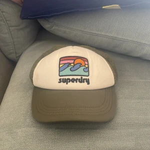 Superdry keps med retrotryck - Snygg keps från Superdry med beige frontpanel och olivgrön skärm. Framsidan har ett färgglatt retrotryck med solnedgång och vågor samt Superdry-logga. Baksidan är i mesh för extra ventilation och justerbar passform. Betala med swish tack!