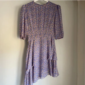 Ljuslila adoore klänning  - Superfin ljuslila klänning med blommigt mönster och volangdetaljer. Klänningen har puffiga korta ärmar och en flowy kjol med lager. Perfekt för vår och sommar!