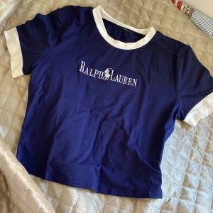 Blå croppad t-shirt från Ralph Lauren - Snygg blå croppad t-shirt från Ralph Lauren med vit logga broderad på bröstet. T-shirten har vita detaljer vid ärmslut och halsringning. Perfekt för en avslappnad och trendig stil.