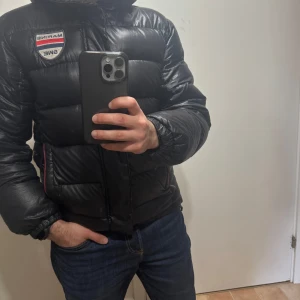 Svart pufferjacka med patch från Marine SWE - Svart pufferjacka från Marine SWE med broderad patch på bröstet. Jackan har dragkedja framtill och röda detaljer längs sidorna. Perfekt för kyligare dagar och har en klassisk pufferlook.