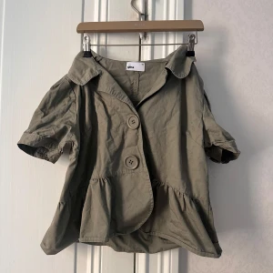 Beige kortärmad ”jacka” med stora knappar från Gina - Säljer en beige ”jacka” från Gina med korta puffärmar och två stora knappar framtill. Jackan har en lös passform och en bred krage, samt volangliknande nederkant. Perfekt för en avslappnad och trendig look. Storleken är 42 men sitter mer som en M💗
