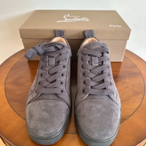 Grå mocka sneakers från Christian Louboutin - Snygga grå sneakers i mocka från Christian Louboutin med klassisk röd sula. Skorna har rund tå, platt sula och snörning. Perfekta för dig som vill ha en stilren och exklusiv look.